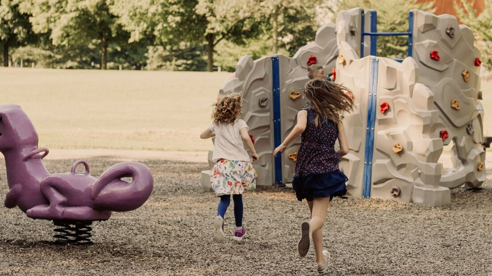 Here are two girls running in the playground of Pinafore Park. The one girl is wearing a colourful skirt, with navy blue leggings and a white top. The other girl is wearing a navy blue and red patterned dressed. they are running towards the rock climbing station of the park.