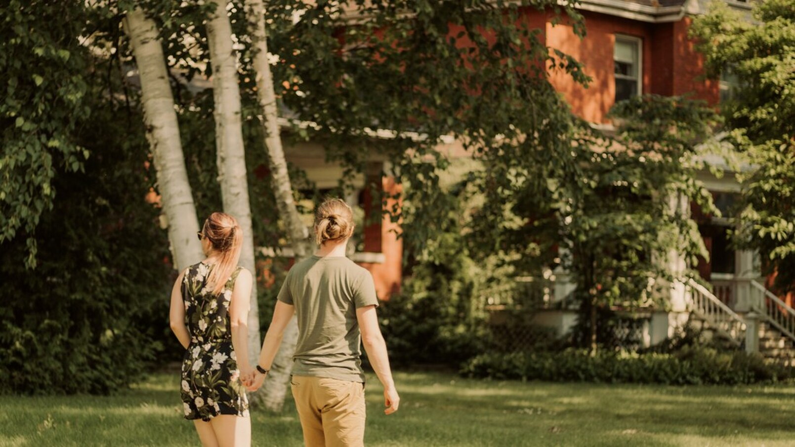 Here is a couple walking in the courthouse district! On the left is a female wearing a floral romper with a black background. She is holding the hand of a man wearing a green shirt and khaki bottoms. All around them is green grass and some trees. Beside them is a red brick house out of focus.