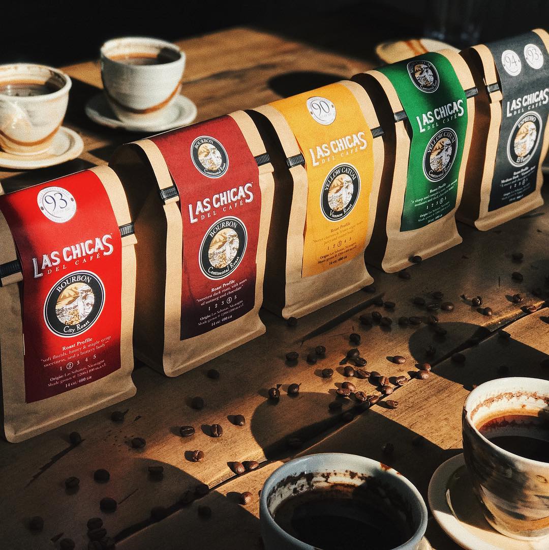 Multiple Las Chicas coffee packages. The first is with an orange label, second is with a red label, the third is with a yellow label, the fourth is with a green label and the fifth is with a grey label. In front and behind the coffee packages are two coffee cups with coffee inside. There are coffee beans spread out on the table.