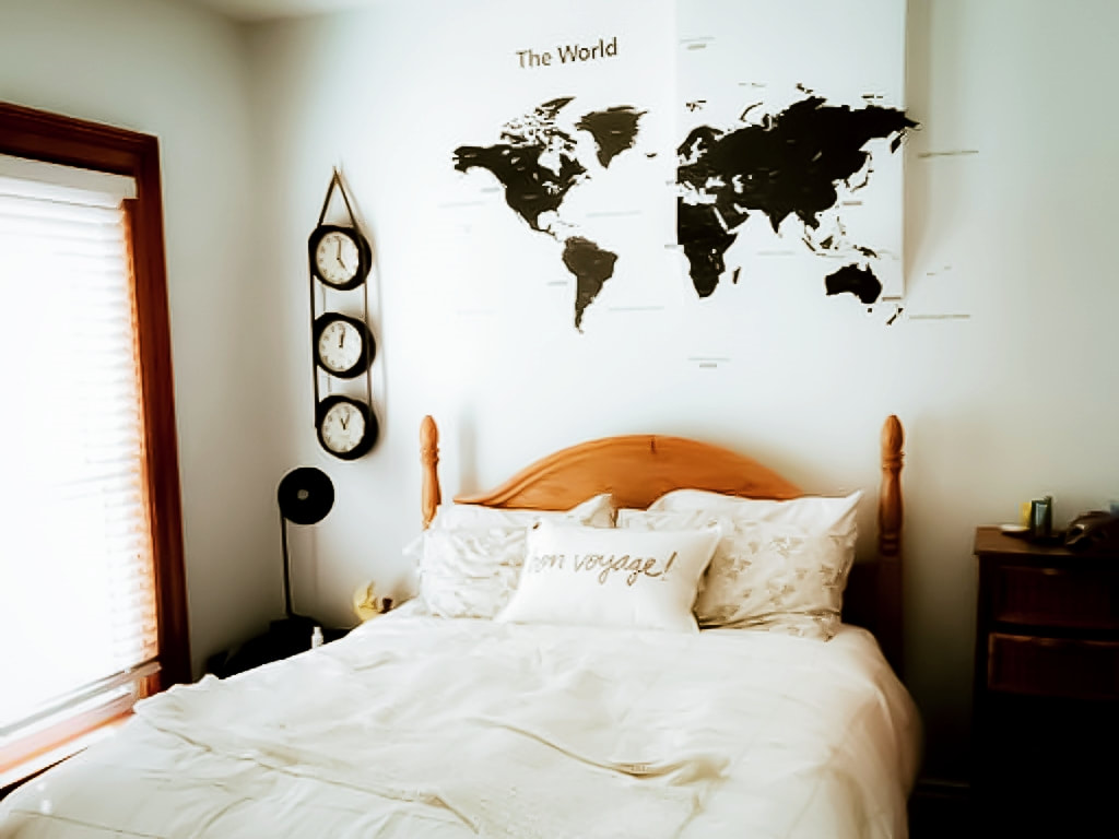 Here is a picture of a bedroom. On the left is a window with white blinds. In the middle us a double bed with a white duvet cover and blanket folded at the end. Above the bed is a world map and three clock handing vertically.