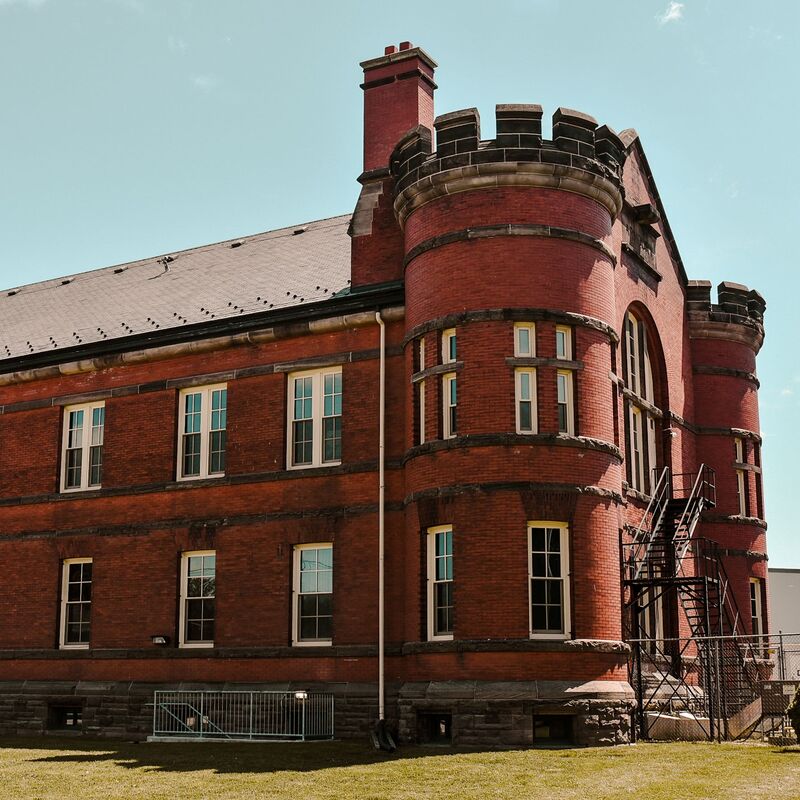 Here is a side angle of the St. Thomas Armoury.  The building is made of red brick with two rows of windows. In the front corners are large round towers. In the front of the middle is a staircase up to the second floor.