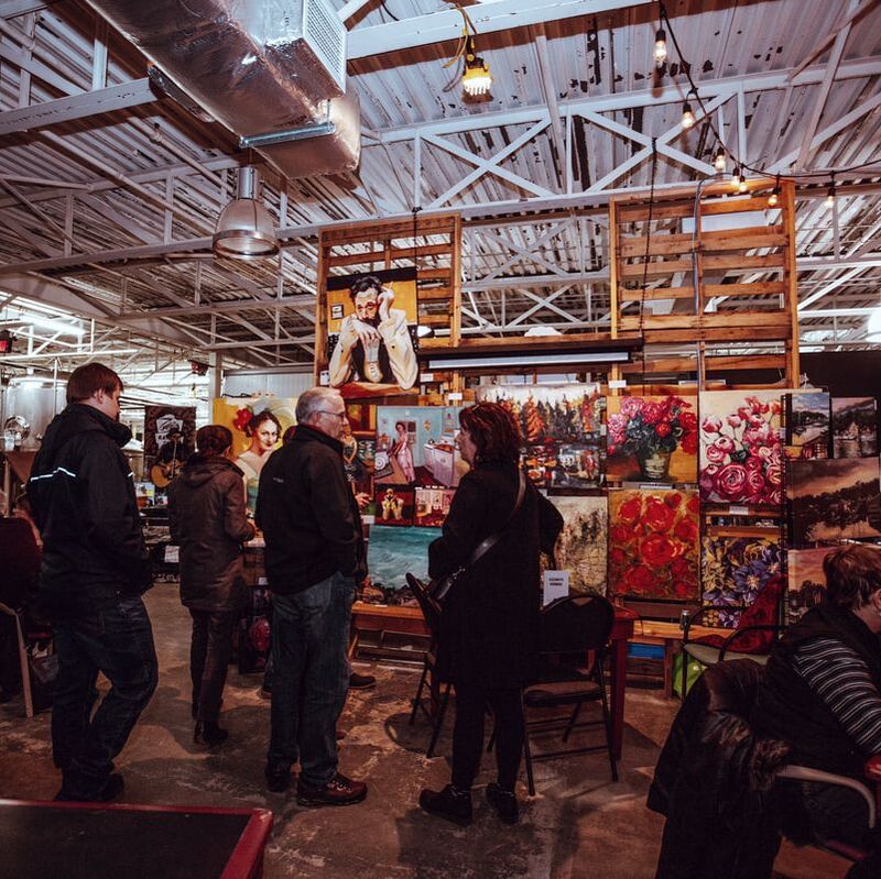This image captures one of the exhibits for the Art Crawl. It is taking place in a warehouse environment, so the ceilings are metal with metal supports painted white. The art exhibit has wood wood pallets hanging from the ceiling to hold up numerous paintings. The first rack displays floral themed and lake landscape paintings. The display on the left shows more portraits of individuals. There are approximately four people in front of the exhibit looking at the art.