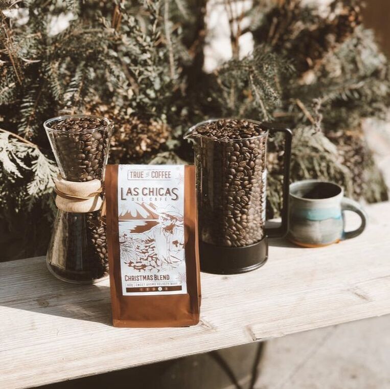 Here is a picture of Las Chicas Del Cafe Christmas Blend. It is in a warm brown bag. Behind the bag are a French press and tall narrow glass filled with coffee beans. Behind the display are pine trees.