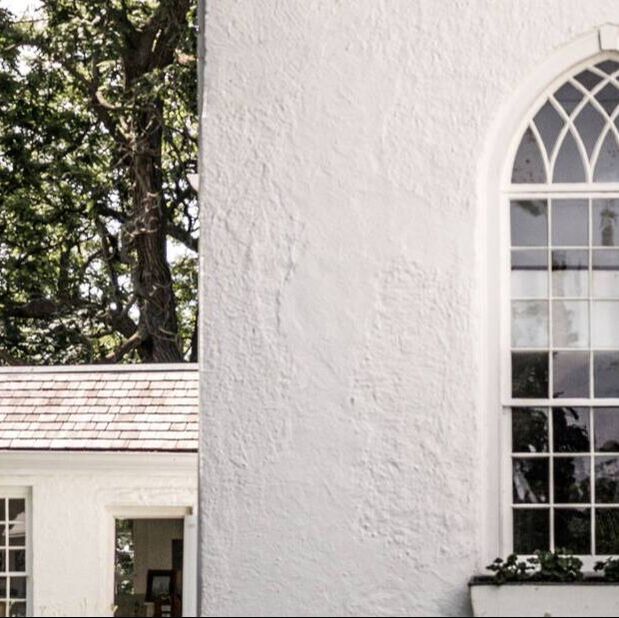 Here is Old St. Thomas Church. This photo shows off one of the corners of the church. The building is completely white. The windows are white paned arched windows. Below the window is a flower box with green plants. To the left of the image is a door which is open leading inside the church.