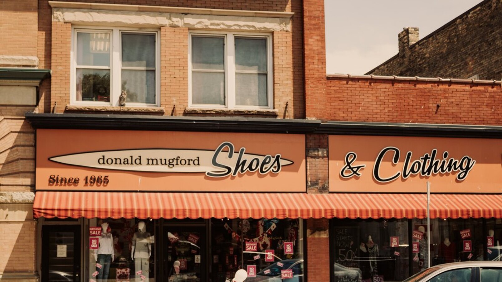 This is the store front of Mugford's. There is an orange and red awning above the store windows. The sign has an orange background and in the bottom left corner of it, it says 