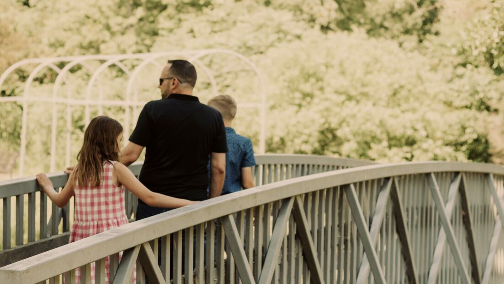 Here is a man and two children crossing one of the bridges at Waterworks Park. The girl is wearing a white and red checkered dress. The man is wearing a black shirt with sunglasses. In front is a boy wearing a blue and white patterned shirt. In the back are many trees.