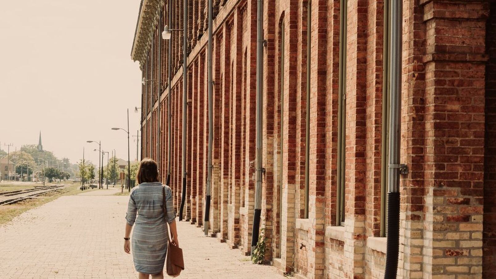 Here is a lady walking beside the CASO Station. The CASO Station is covered in red brick and each window has an olive green frame. The lady is walking with a brown purse on her shoulder and is wearing a blue and white dress. To the left of the shot are some train tracks.