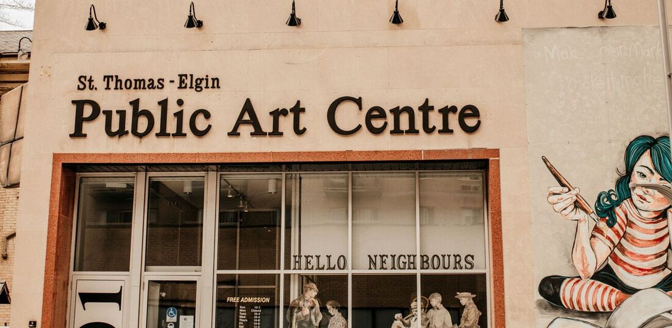 Here is the front of the arts centre. On the right is a mural with a little girl with blue hair and a paint brush. Straight ahead are windows with people drawn on them. To the left of the windows are a glass door into the gallery,