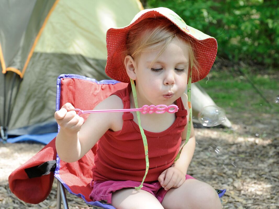 Here is a little girl wearing a red tank top and sunhat. She is sitting in a red and blue foldable chair with a green and yellow tent behind her. She is leaning forward and blowing out bubbles.
