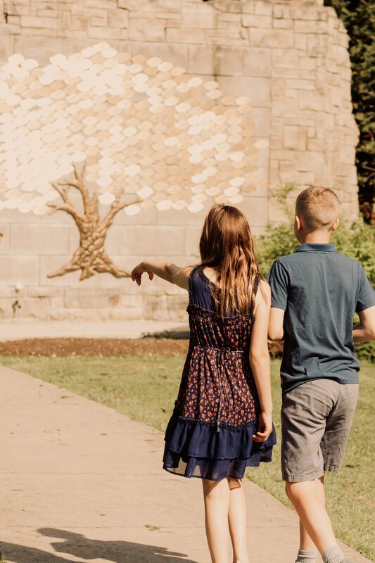A girl and boy walking on a path in Pinafore Park. The girl is wearing a navy blue dress with red flowers. The boy is wearing beige khaki pants and a navy blue top. They are pointing to the memorial tree on the wall in front of them. In front of the wall is mulch and then grass.