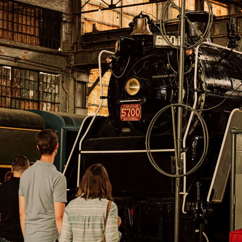 Here is an image of a black train. There is a red license plate in the middle with the numbers “5700”. There is a woman with brown hair and a blue and white striped shirt as well as a man with brown hair and a light blue shirt. Behind him is another man wearing a black shirt. 