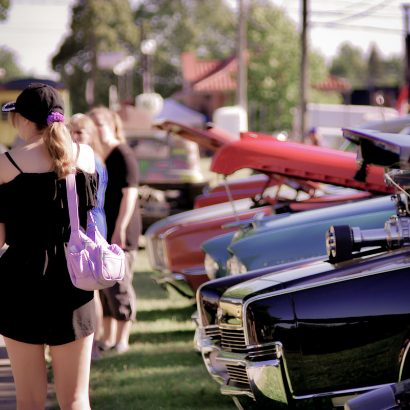 There is a person walking away from the camera. She is wearing a black romper and ball cap and has a purple purse and scrunchie. Beside her on the right are old fashion cars. The first one is a dark plum purple, then a periwinkle, and the last two cars are red.