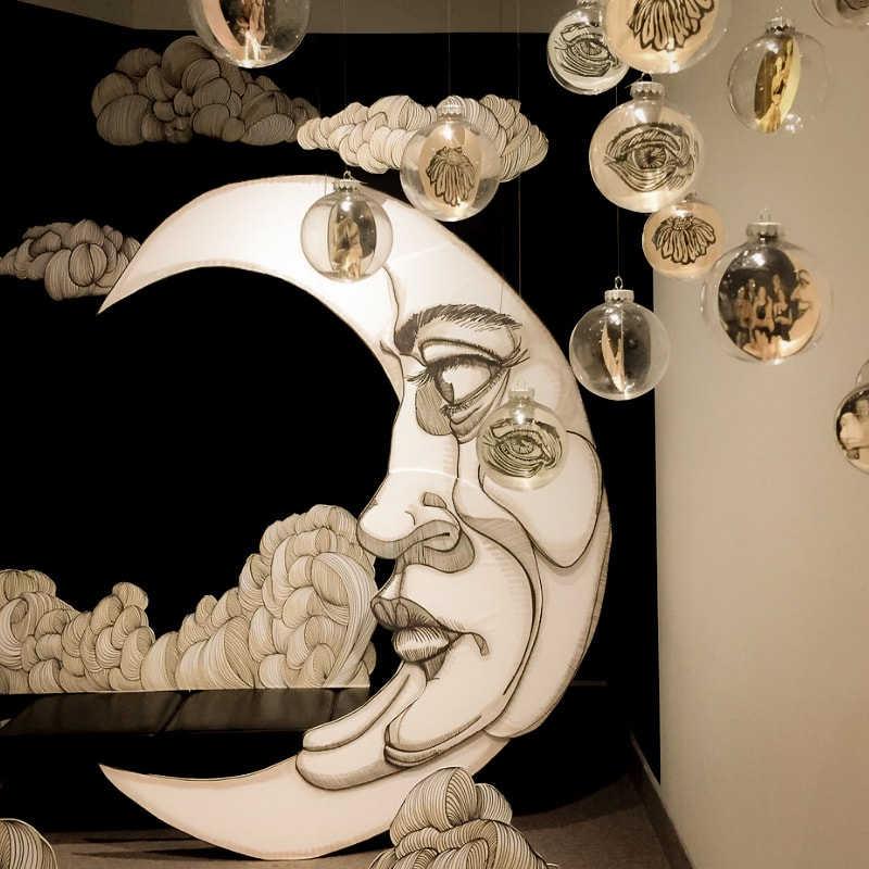 Here is an image of an art exhibit at the St. Thomas Public Art Gallery. In the centre is an image of a crescent moon. The moon has an eye brow, eye, nose and mouth. Behind the moon are cloud bigures and in front are glass ornaments with photos inside.